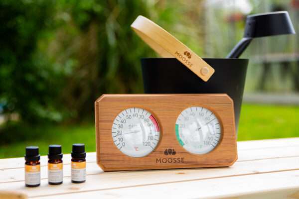 MOOSSE sauna thermometer naast sauna aroma geuren, en op de achtergrond een MOOSSE aluminium sauna emmer met lepel.