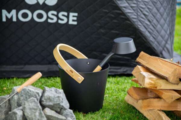 Aluminum MOOSSE sauna emmer waar water in wordt gegiet, naast saunastenen en kachelhout en een vuurpoker.