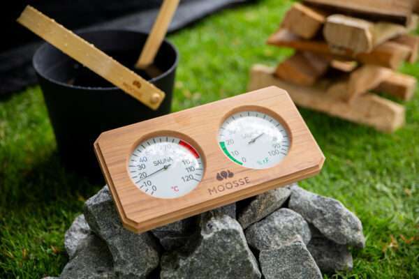 MOOSSE thermometer op een berg saunastenen, met op de achtergrond een aluminium MOOSSE sauna emmer met lepel en kachelhout.